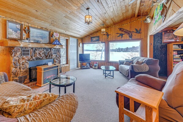 A cozy cabin interior with a stone fireplace, wooden walls, a television, and a large window overlooking a snowy landscape. - Lakefront Suring Home w/ Pool Table + Views! (Suring)