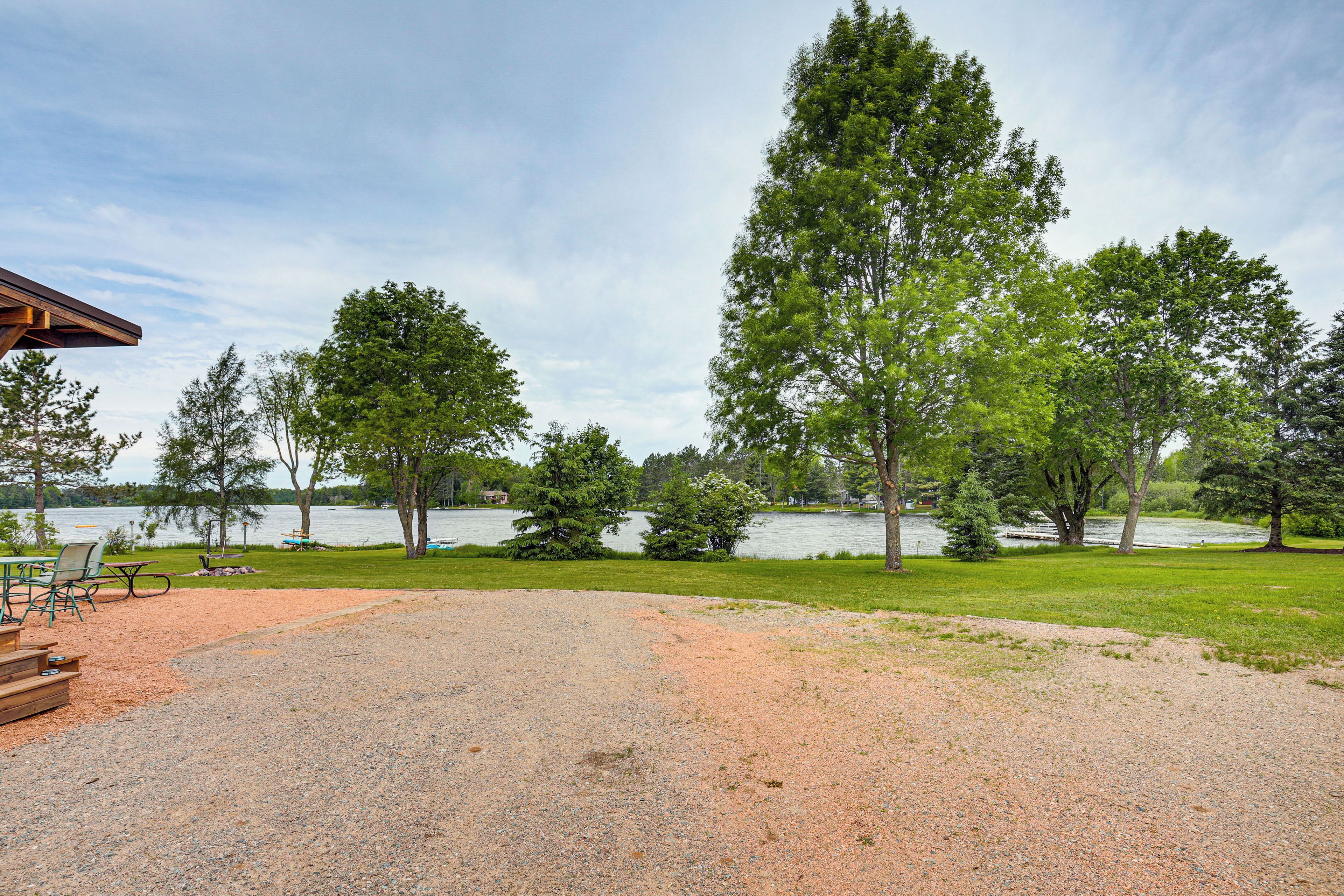 A gravel area with picnic tables and a wooden deck, surrounded by trees and a lake.