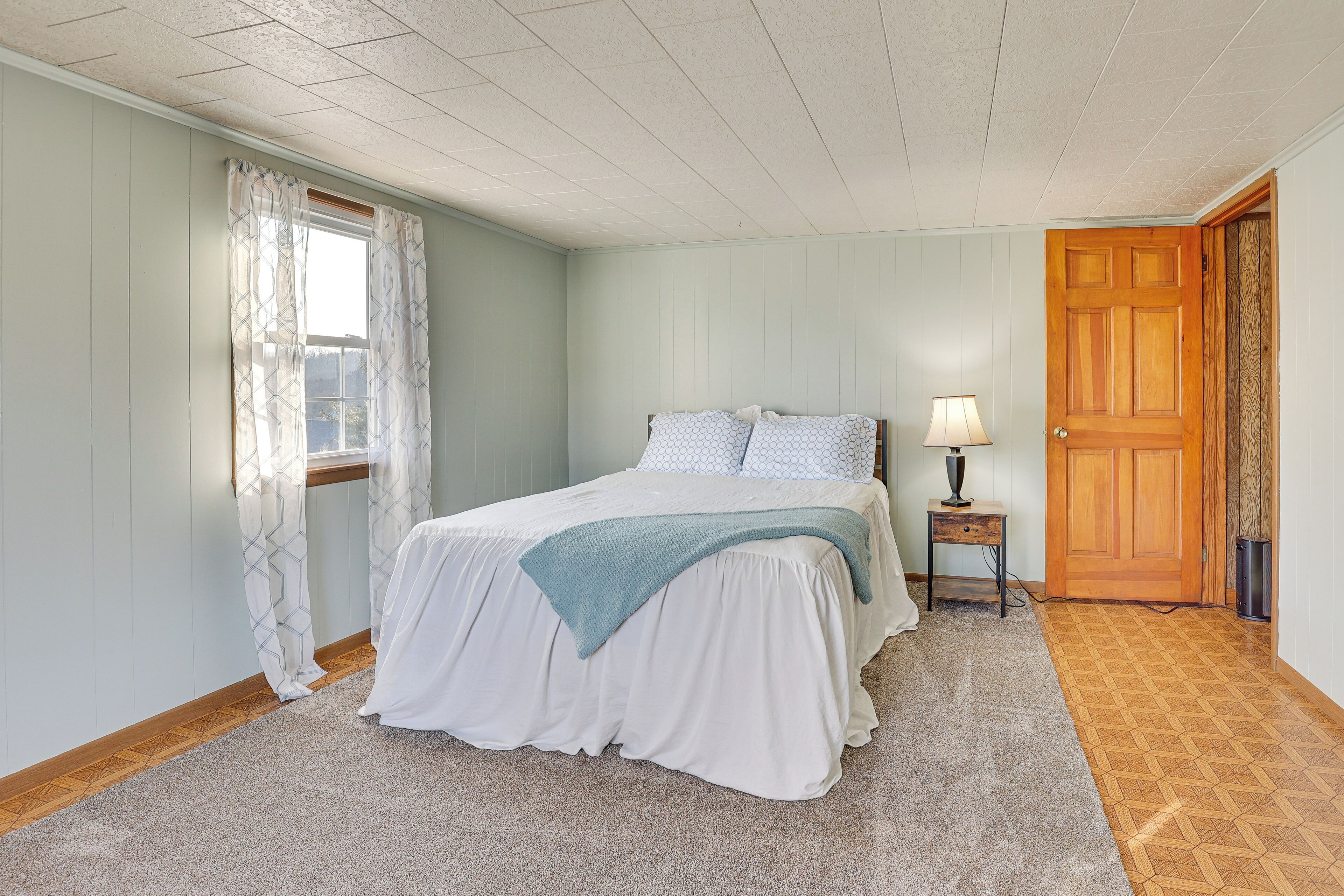A bedroom with a bed, a nightstand, a lamp, a window with curtains, and a wooden door.