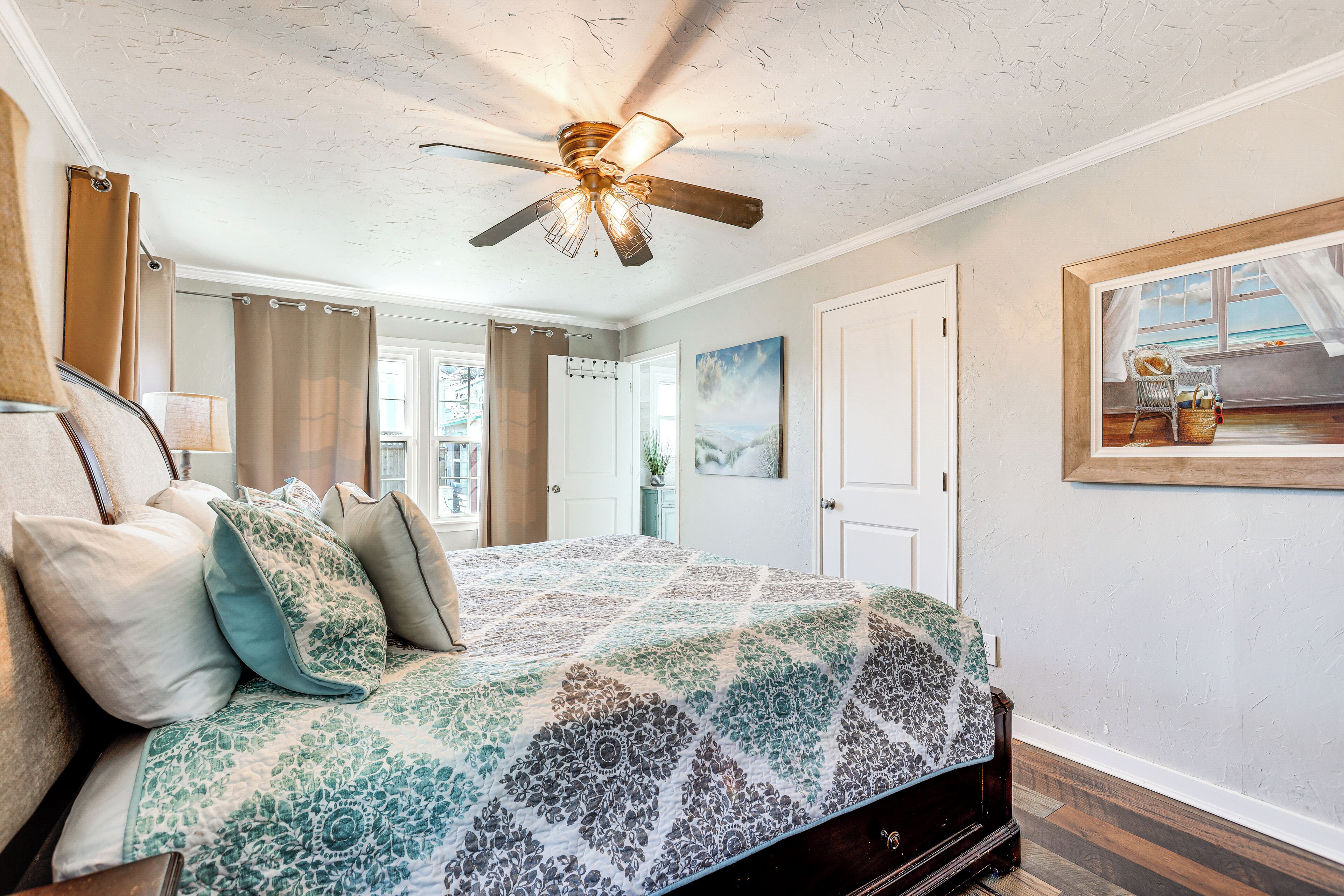 A bedroom with a bed, a ceiling fan, a painting, and a view of the outdoors.