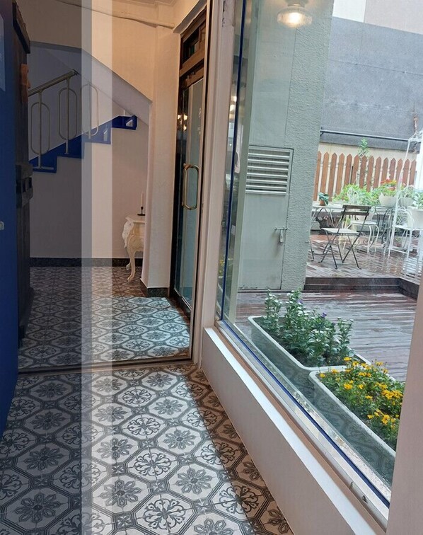A narrow hallway with patterned floor tiles, a glass door leading outside, and a view of a patio with outdoor furniture and plants.