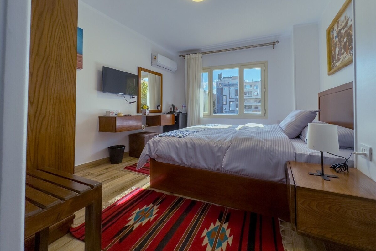 A bedroom with a bed, a TV, a desk, and a window with a city view.