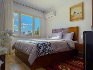 A hotel room with a bed, a window with a view, a wall-mounted air conditioner, and a framed picture. - Royal Crown Hotel Giza Pyramids View (Giza)
