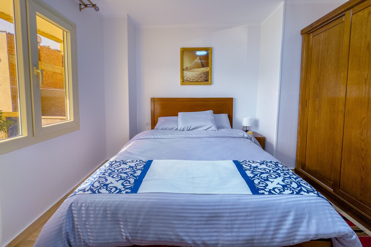 A bedroom with a bed, a nightstand, a wardrobe, a window, and a painting on the wall.
