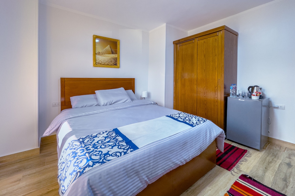 A neatly made bed with a patterned bedspread, a wooden wardrobe, and a small kitchenette area with a kettle and glasses.