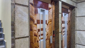 Un ascensor con diseño de pared de piedra, una lámpara moderna y un botón de salida de emergencia visible.