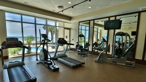 Une salle de sport équipée de tapis de course, de vélos elliptiques et d’appareils de musculation. De grandes fenêtres offrent une vue sur l’océan.