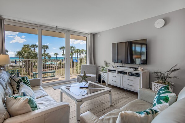 A living room with a TV, sofa, coffee table, and a balcony with a view of palm trees and the ocean.
