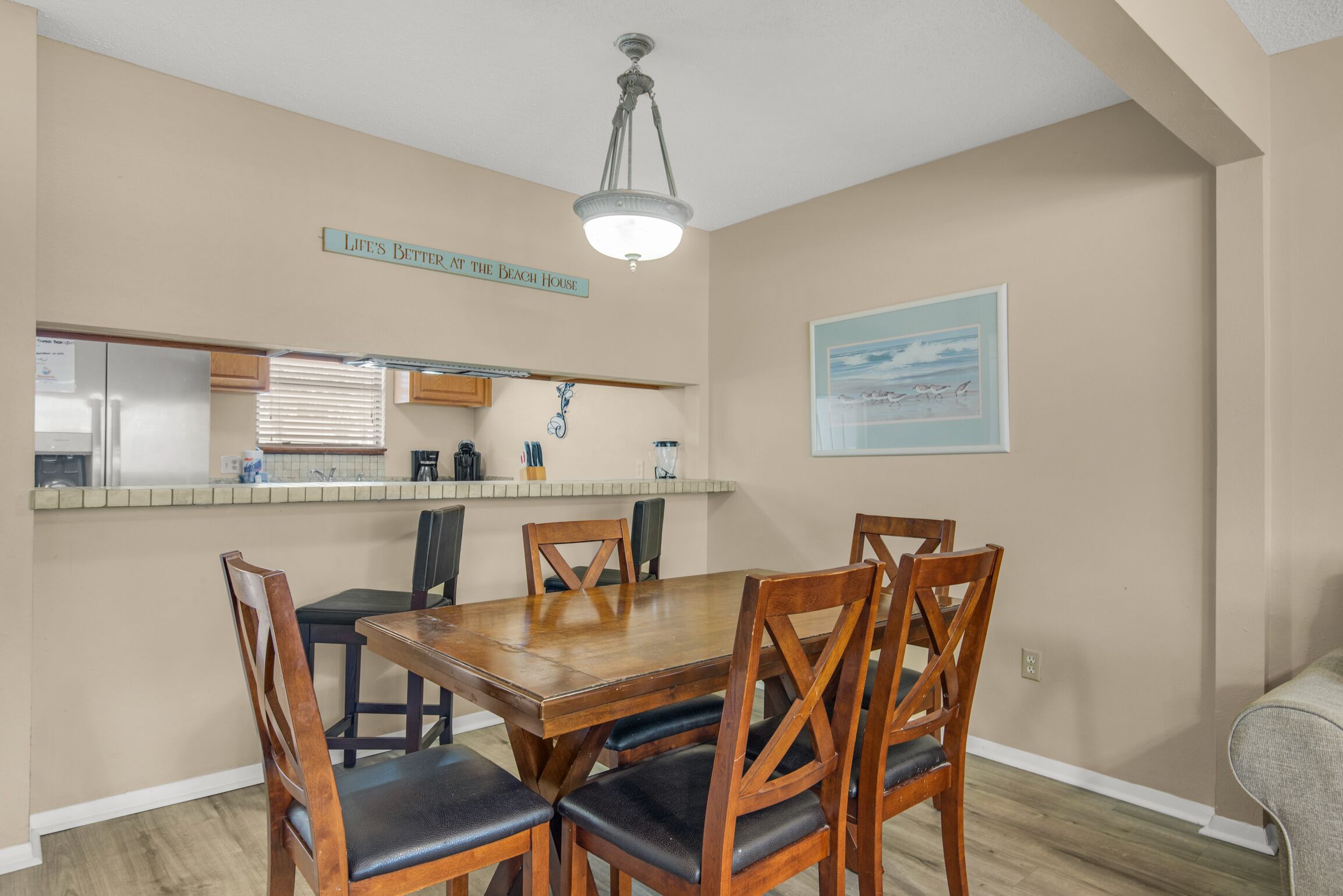 A dining area with a wooden table and chairs, a kitchen with a refrigerator and countertop, and a ceiling light fixture.