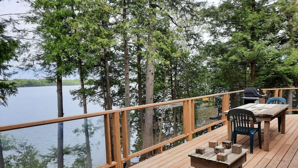 Een houten terras met tafel en stoelen met uitzicht op een meer, omringd door bomen.
