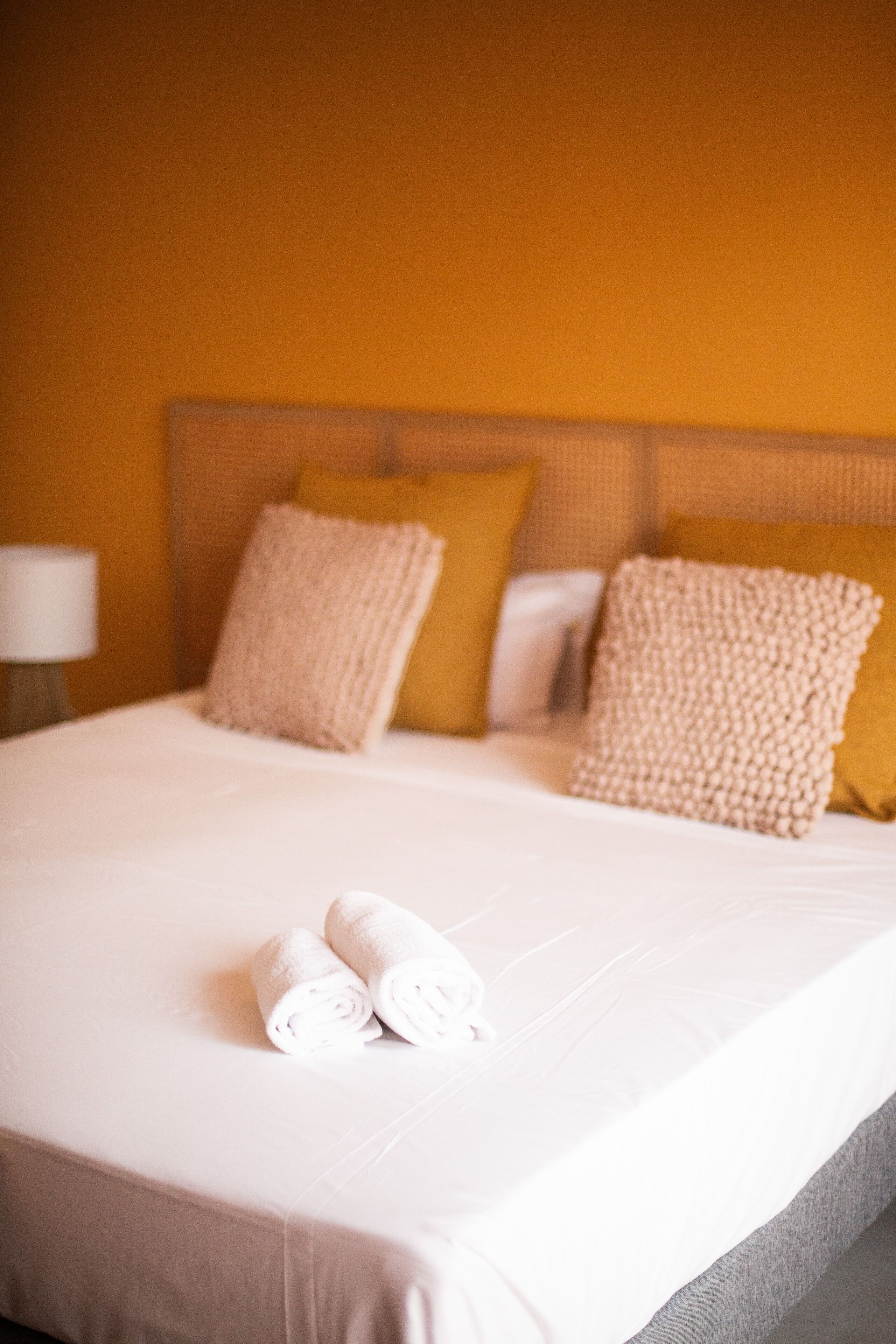 Una cama bien hecha con toallas enrolladas, cabecera con varios almohadones y una lámpara sobre la mesita de noche.