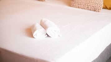 Una cama bien hecha con toallas enrolladas, cabecera con varios almohadones y una lámpara sobre la mesita de noche.