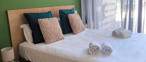 Una cama bien hecha con sábanas blancas y dos toallas enrolladas, un cabecero de madera y una pared verde.