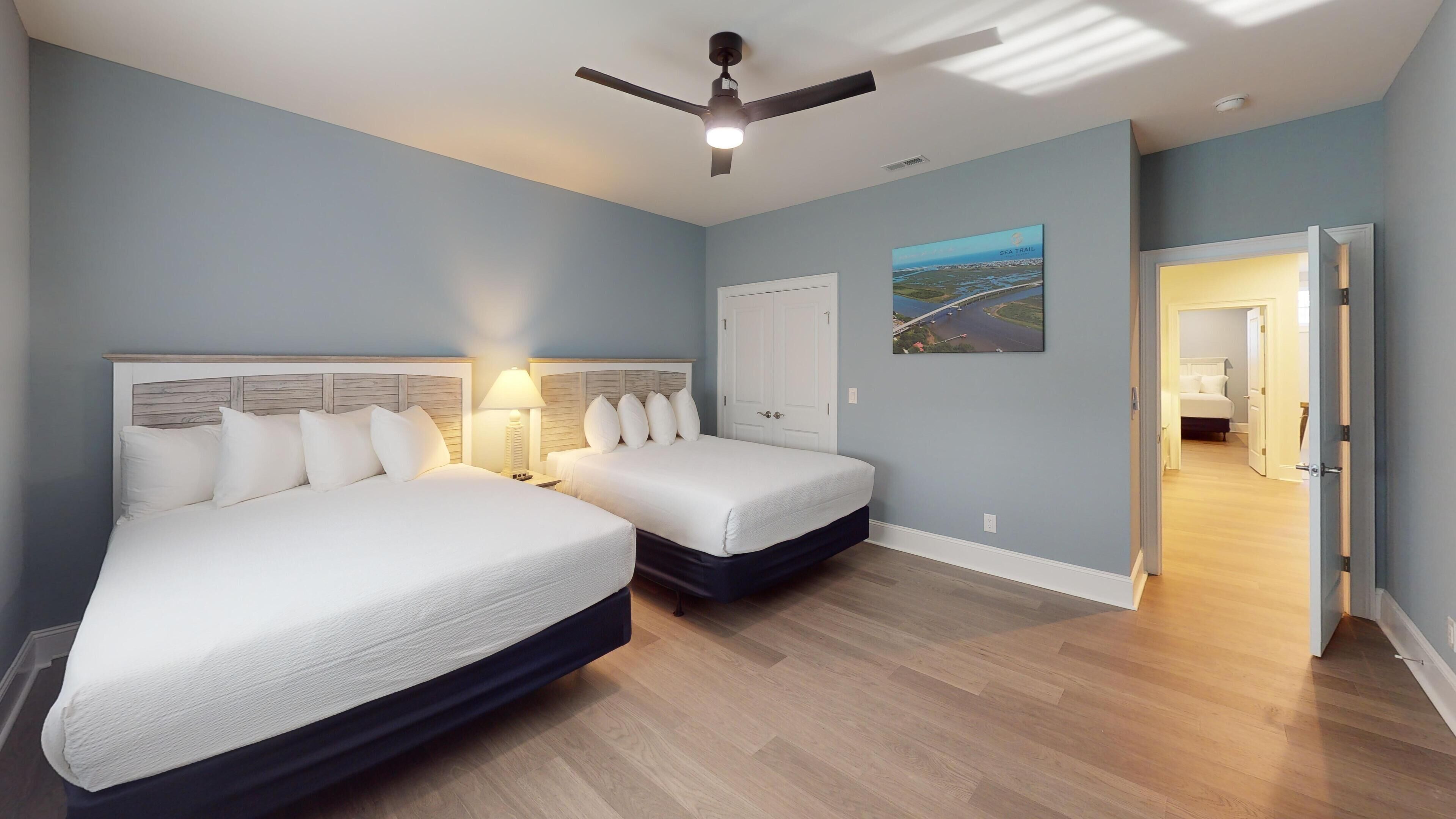 A hotel room with two beds, a ceiling fan, a painting of a bridge, and a view of a room with a bed.