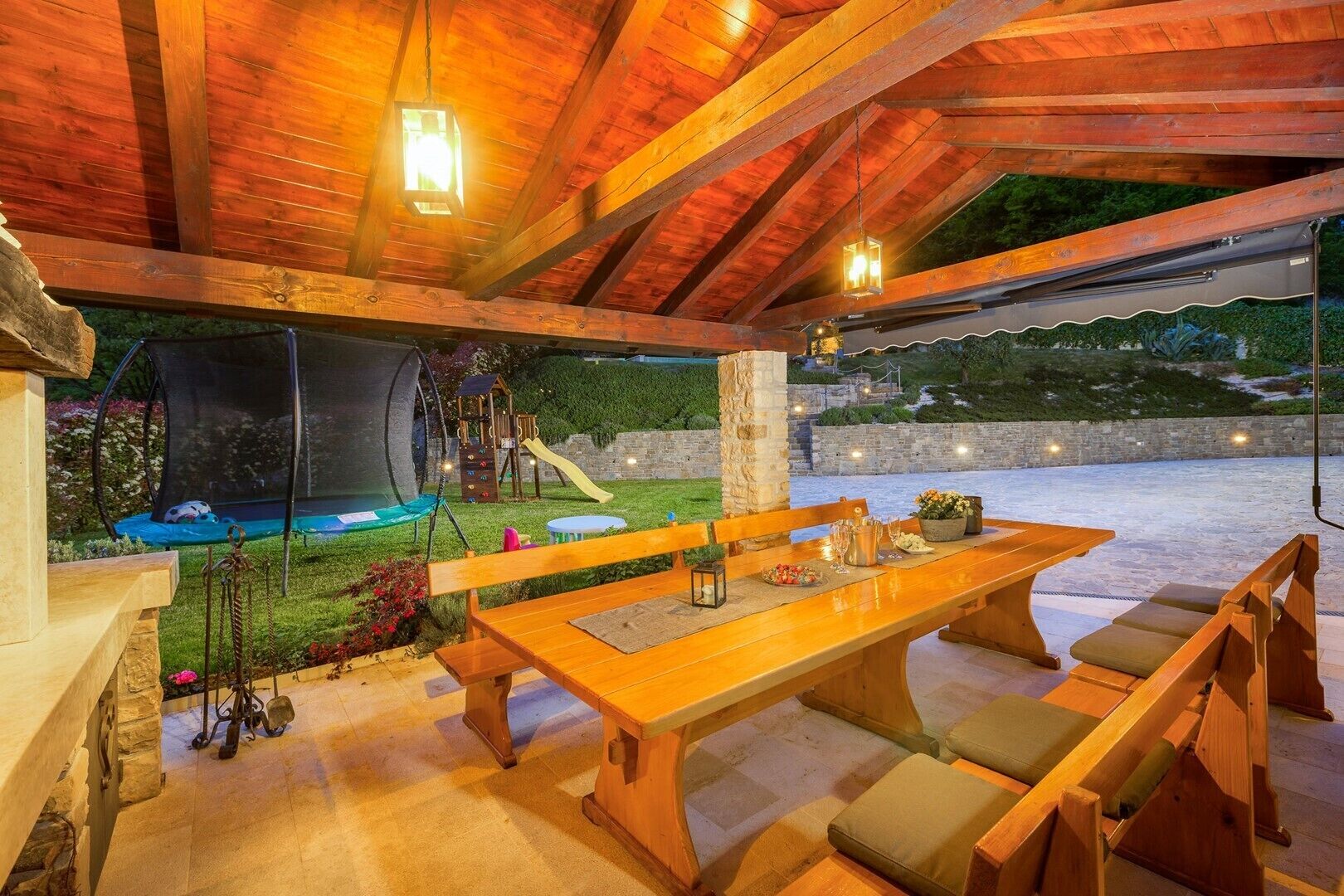 Un espace de restauration en plein air avec une table et des bancs en bois, donnant sur un jardin avec un trampoline et un ensemble de jeux pour enfants.