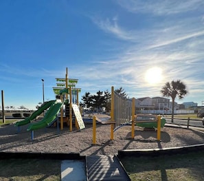 A playground with a slide and climbing structures, surrounded by a fence and a clear sky. - Toes in the Sand - Ocean Views, Steps from Beach (Oak Island)