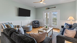 A living room with a ceiling fan, a flat-screen TV, a dark armchair, a round ottoman, and a coffee table with decorative items. - Toes in the Sand - Ocean Views, Steps from Beach (Oak Island)