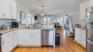 A modern kitchen with white cabinets, a dishwasher, a coffee maker, and a dining area with a chair and table. - Toes in the Sand - Ocean Views, Steps from Beach (Oak Island)