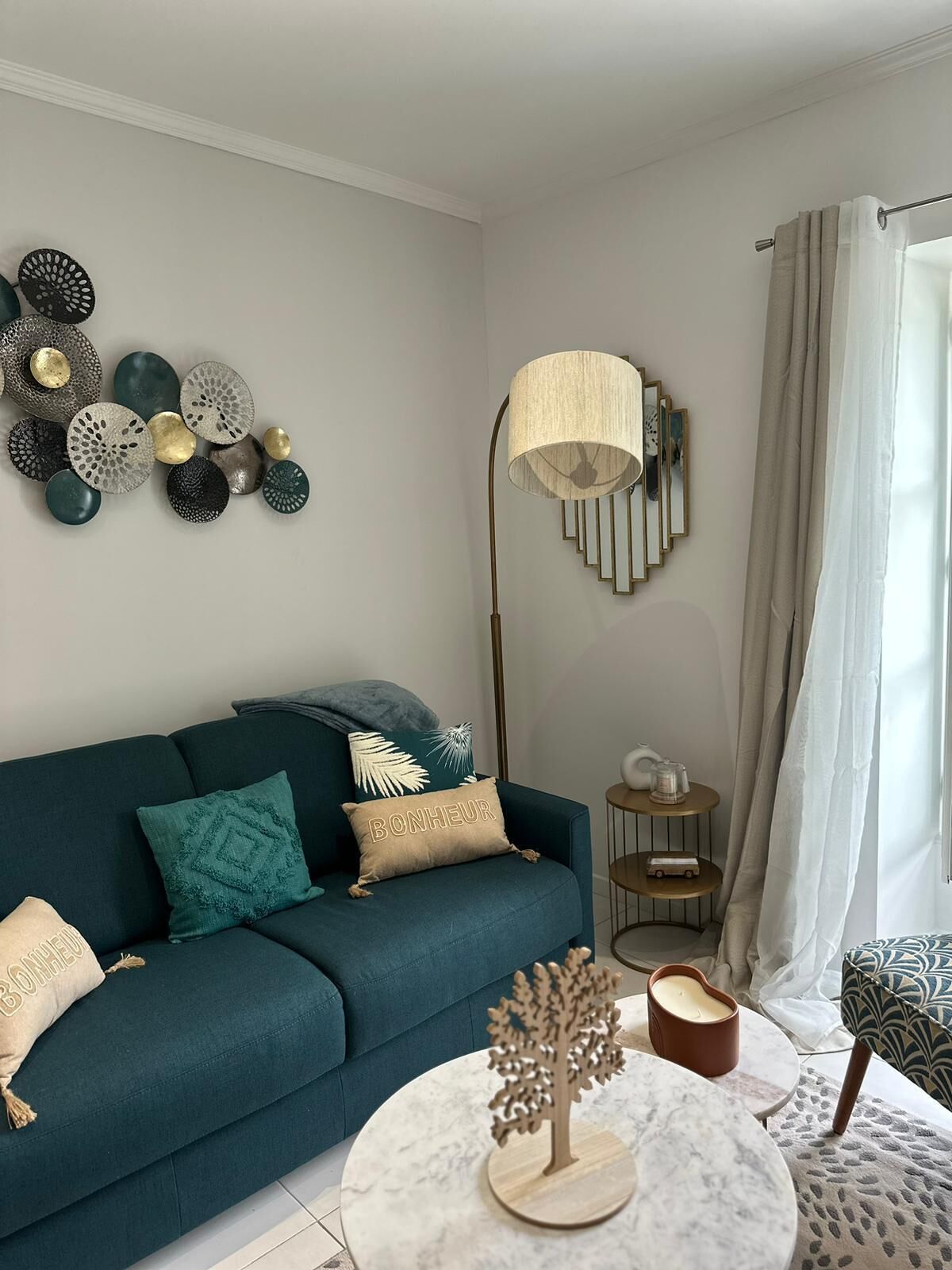 Un salon avec un canapé de couleur turquoise, des œuvres d’art décoratives accrochées au mur, une lampe sur pied et une petite table ronde ornée d’une bougie et d’une décoration en forme d’arbre en bois.