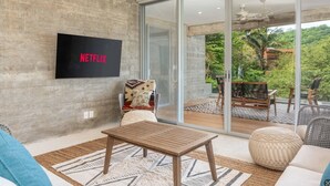 A modern living room with a flat-screen TV displaying "NETFLIX," a large sliding glass door leading to a balcony with outdoor furniture, and a cozy seating area with a patterned rug. - Modern Condo In Walking Distance to Surf Beach (Bahias de Huatulco)