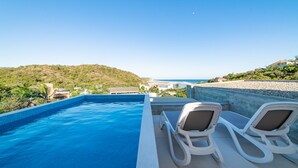 A swimming pool with two lounge chairs and a view of the beach and hills. - Modern Condo In Walking Distance to Surf Beach (Bahias de Huatulco)