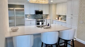 Une cuisine moderne avec des meubles blancs, un îlot central doté de tabourets de bar, un réfrigérateur en acier inoxydable et un lustre.