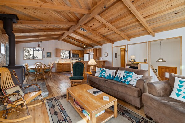 A spacious living room with a wooden ceiling, a brown sofa with patterned cushions, a wooden coffee table, and a rocking chair.