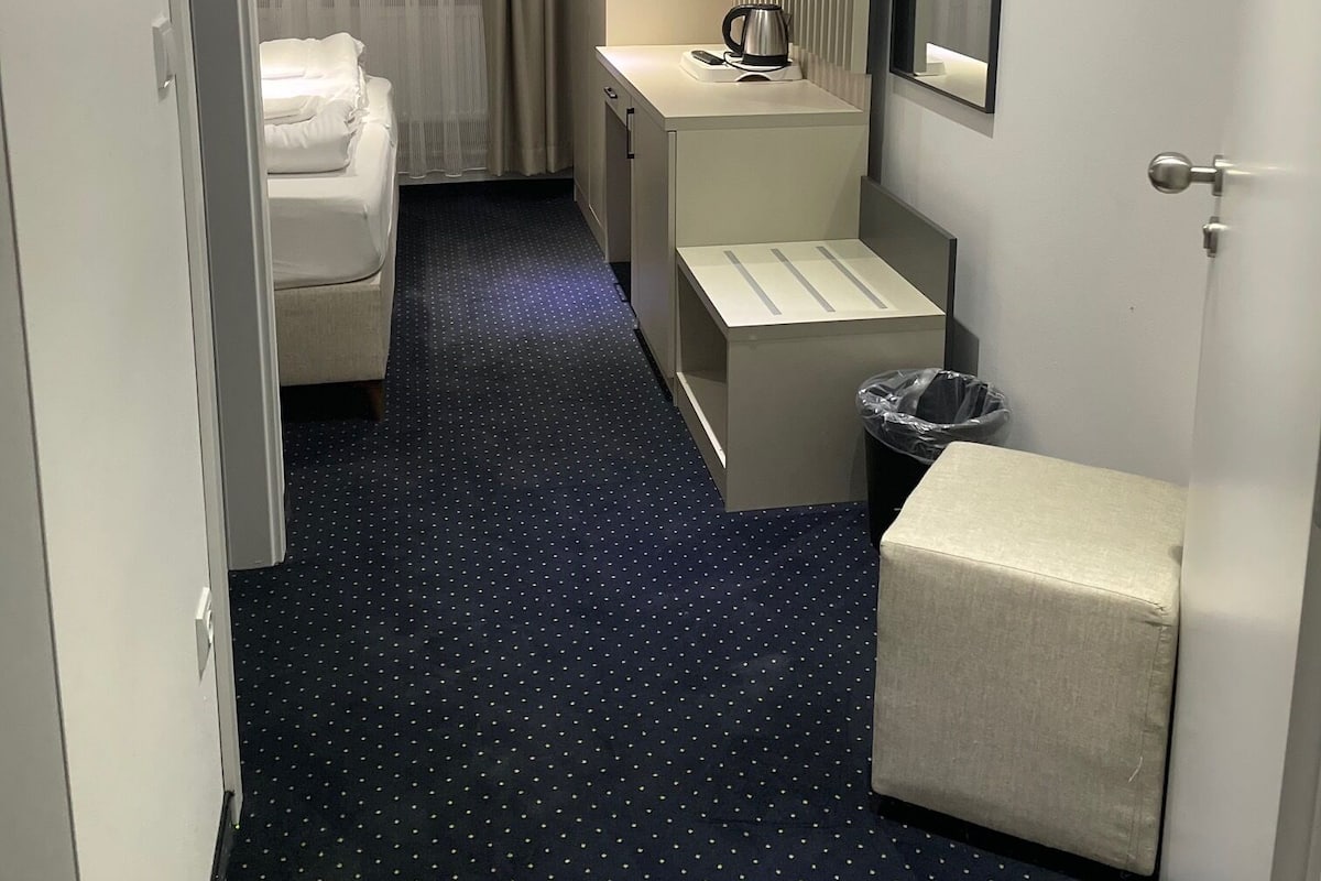 Quarto de hotel com uma cama, uma pequena escrivaninha com chaleira, um cesto de lixo e um espelho.