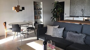En moderne stue med sofa, spisebord med stole, reol og et køkkenområde med vask.