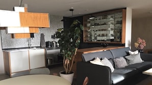 En moderne stue med en sort sofa, et rundt bord og en stor stueplante. Der er et køkkenområde med skabe og en bordplade.