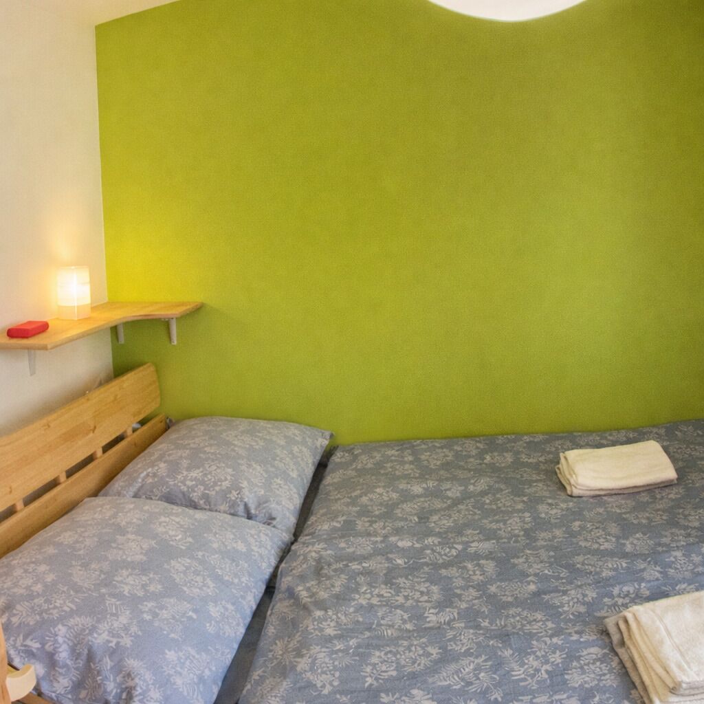 A room with a bed, a wooden headboard, a green wall, and a small shelf with a lamp.
