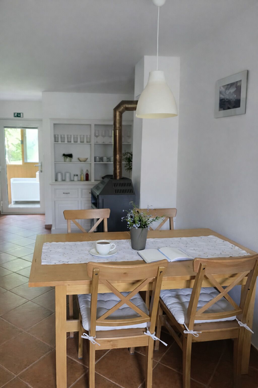 Un comedor con una mesa de madera, cuatro sillas, una lámpara colgante blanca y vista a una cocina con estufa y estantes.
