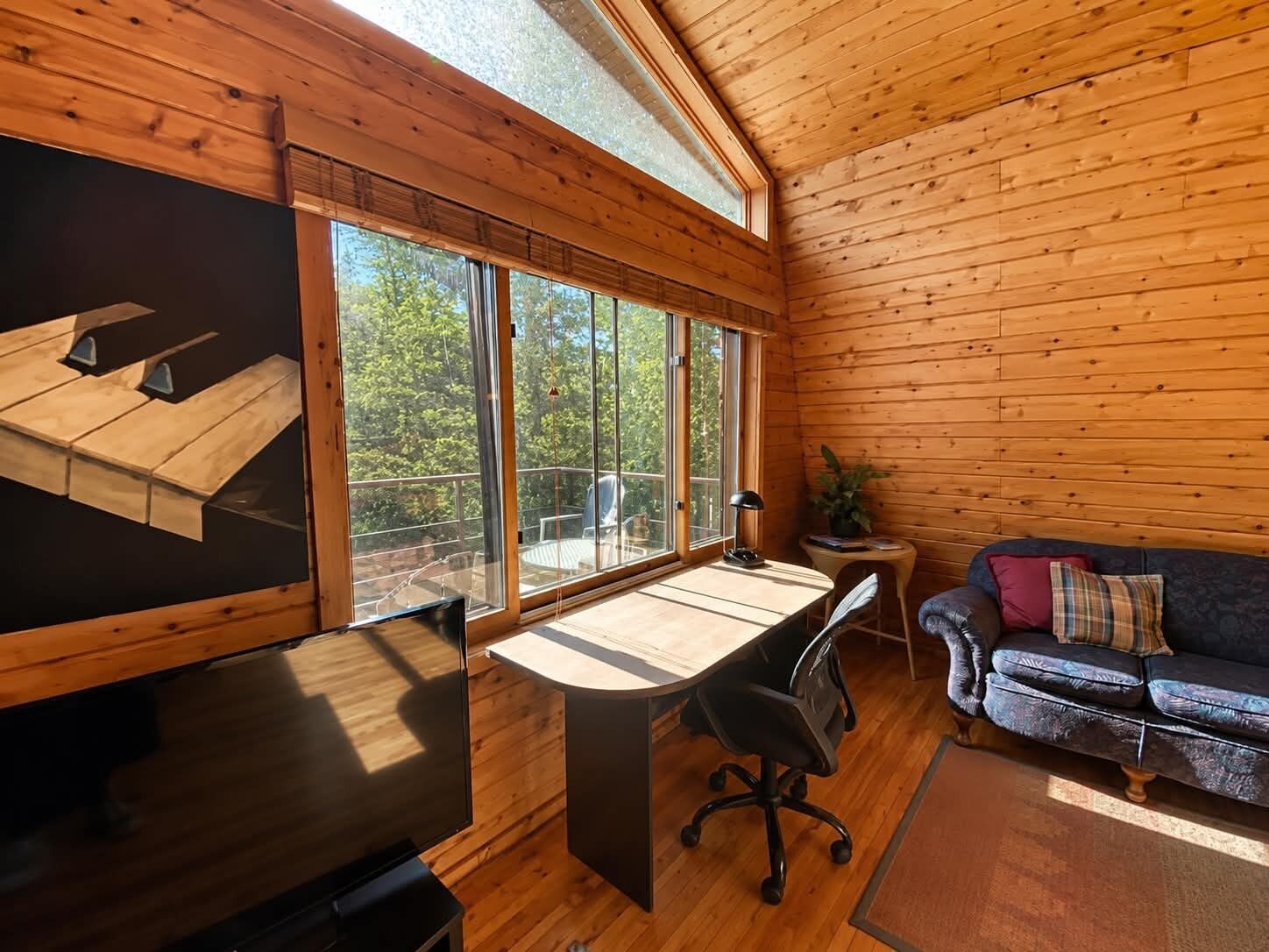 A cabin interior with a wooden interior, a desk with a chair, a sofa, and large windows offering a view of the outdoors.