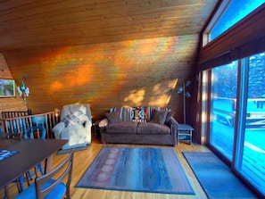 Un interior acogedor de cabaña con techo de madera, un sofá, una mesa de comedor y un ventanal grande.