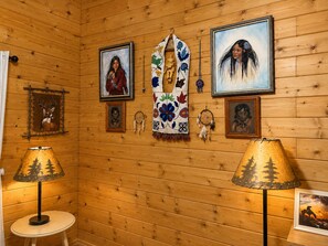 Una habitación con paredes de madera, cuadros enmarcados, un manto tejido en el centro y dos lámparas.
