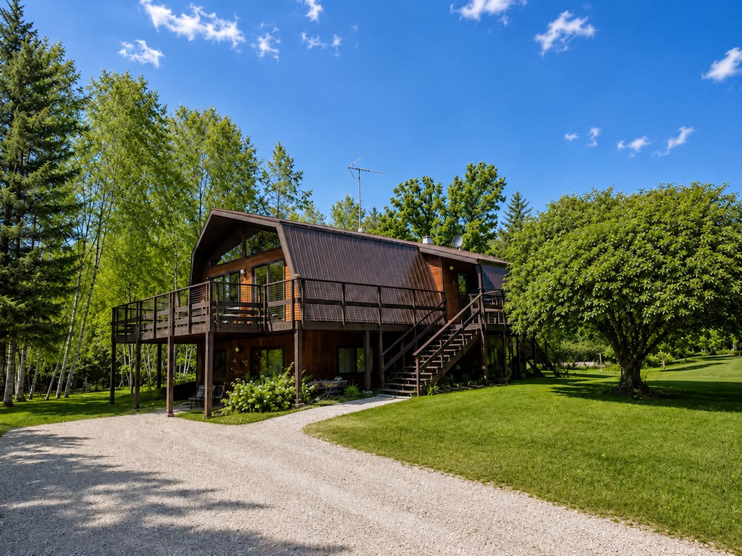 A two-story wooden cabin with a balcony, surrounded by a gravel driveway and lush greenery.