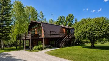 A two-story wooden cabin with a balcony, surrounded by a gravel driveway and lush greenery.