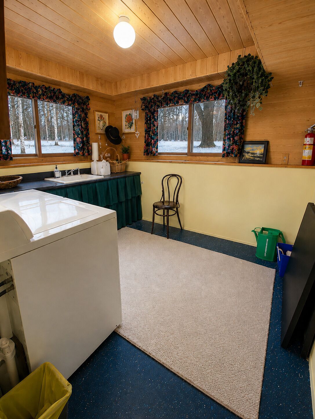 Ein Zimmer mit Waschmaschine, einem Sessel, einem Tisch mit grüner Tischdecke, einem Fenster mit Blick auf Bäume und einem Feuerlöscher.