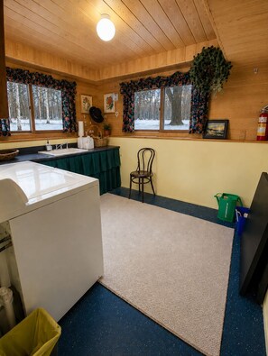 A room with a washing machine, a chair, a table with a green tablecloth, a window with a view of trees, and a fire extinguisher. - 30% OFF Members. Riverfront. Lake Of the Woods 2mi. Pets Free (Warroad)