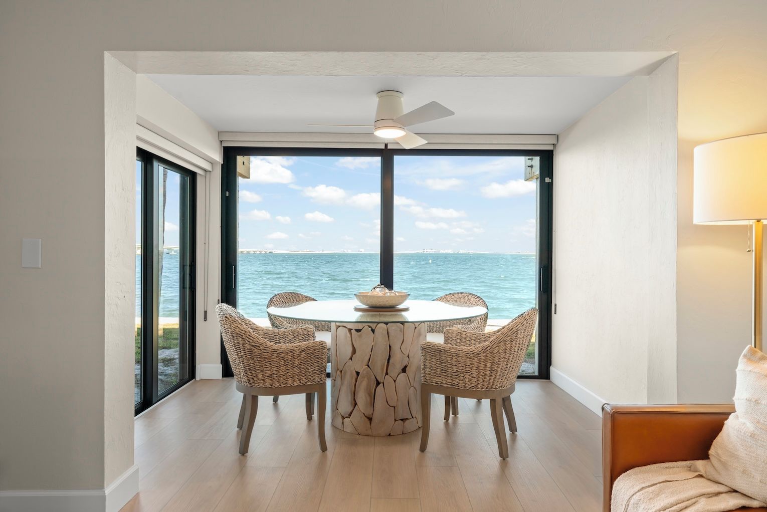 A dining area with a round table and wicker chairs, large windows with a view of the sea, and a ceiling fan.