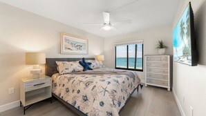 A bedroom with a bed, nightstands, a ceiling fan, a window with a view of the sea, a dresser, and a TV mounted on the wall. - Mariner Pointe #1021 – Sanibel Waterfront! (Sanibel)