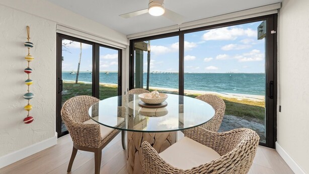 A dining area with a glass table and wicker chairs, overlooking a beach with boats and a clear sky. - Mariner Pointe #1021 – Sanibel Waterfront! (Sanibel)