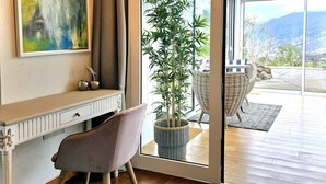 Een kamer met een bureau, een stoel, een grote potplant en uitzicht op de omgeving via een glazen deur.
