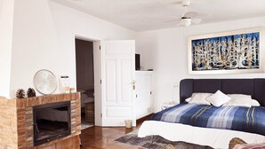 Een slaapkamer met een open haard, een bed met een blauw-wit gestreepte dekbedovertrek, een groot abstract schilderij aan de muur en een plafondventilator.