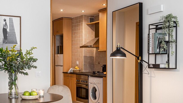 Een moderne hotelkamer met een bed, een eettafel met fruit en een boek, een keukenhoek met een wasmachine en een zithoek met een lamp en een plant.