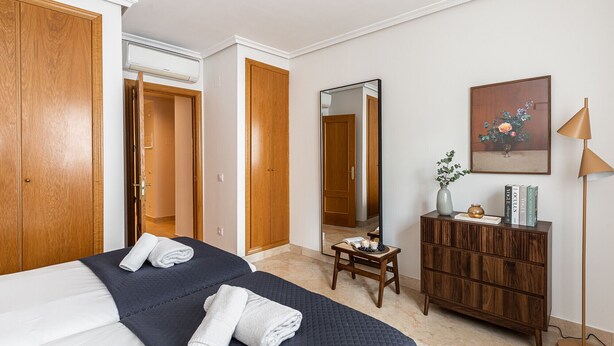 A hotel room with two beds, a wooden wardrobe, a mirror, a small table with a vase, and a floor lamp. - Waou Gran Via Apartment IV - Corta Duració (Madrid)