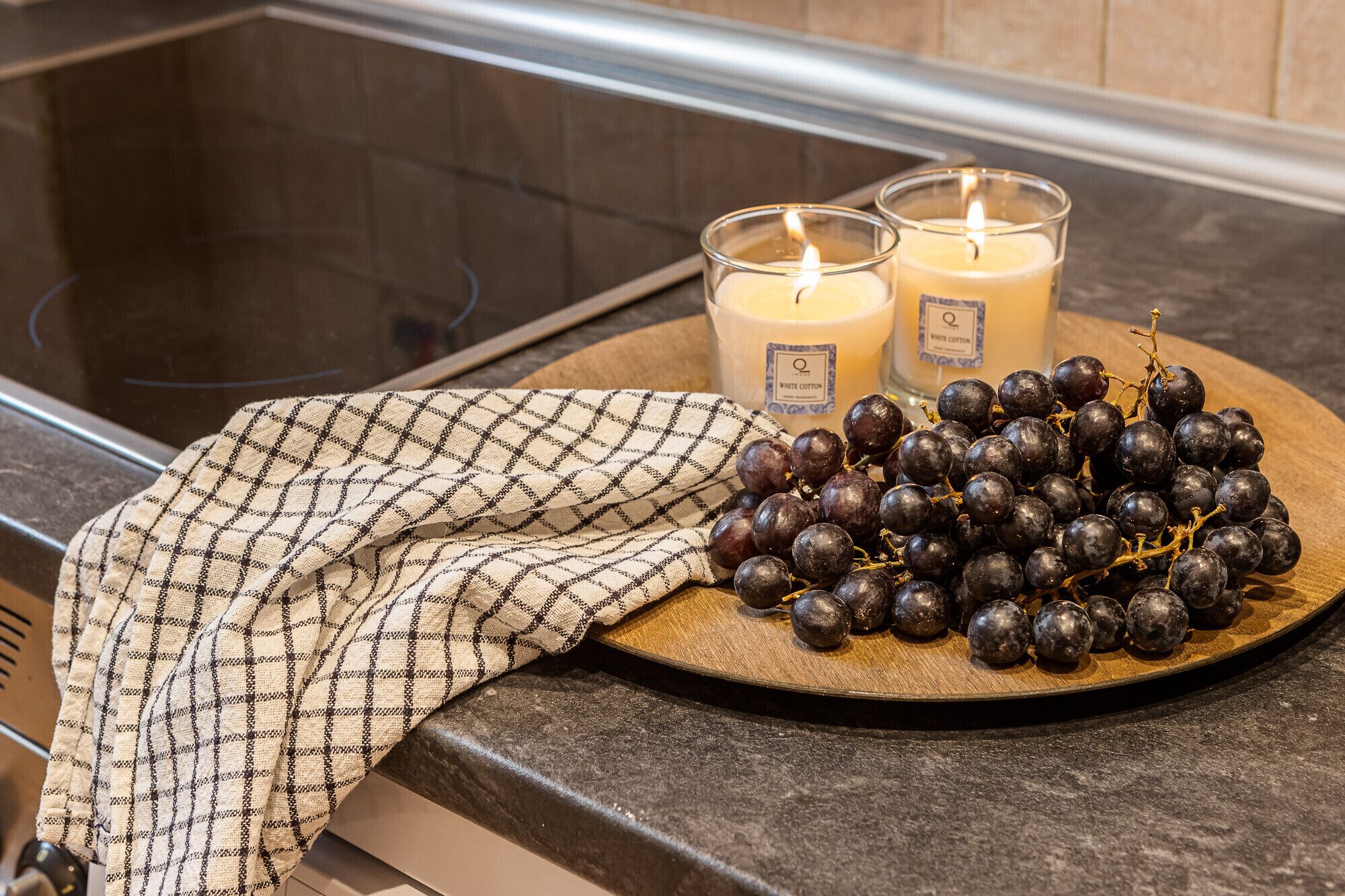 A kitchen counter with a checkered cloth, two lit candles, and a plate of dark grapes.