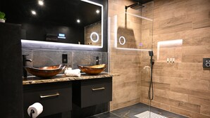 A modern bathroom with a large mirror, two sinks with copper bowls, a black vanity, a shower with a glass door, and a marble floor.