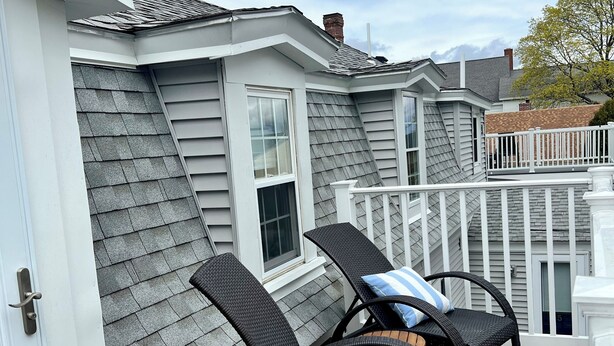 A covered outdoor deck with two wicker chairs and a small table, white railings, and a grey shingled roof. - The Ogunquit Getaway - 177 Shore Road (Ogunquit)