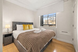 Ein modernes Schlafzimmer mit einem großen Bett, einem Nachttisch, einem Fenster mit Blick auf Gebäude und einer Tür.